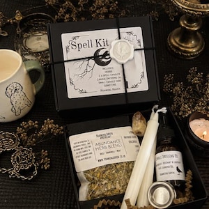 Könnte beinhalten: Eine schwarze Schachtel mit der Aufschrift Spell Kit und einem weißen Wachssiegel, die einen Beutel mit Abundance Kräutermischung, zwei weiße Stabkerzen, eine kleine Flasche Kerzenöl, einen Metallkerzenhalter und einen Edelstein enthält.