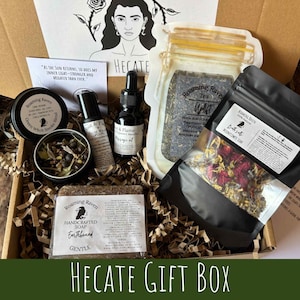 May include: A Hecate gift box containing a selection of items. It features a jar of tea, a black bottle of massage oil, a tin of Green Witch Smoke Blend, a bar of Earthbound soap, and a bag of Moonflower Soak. The box has a drawing of a woman and the text "Hecate Gift Box".