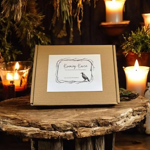 May include: A brown cardboard box with a white label featuring the text "Roaming Raven" and a raven illustration. The box is on a rustic wooden surface, surrounded by candles and greenery, creating a mystical ambiance.