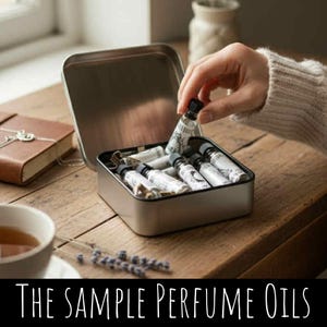 Puede incluir: Una caja metálica que contiene varios frascos pequeños de aceites de perfume con etiquetas blancas y texto negro. Una mano sostiene uno de los frascos. El texto "THE SAMPLE PERFUME OILS" está en la parte inferior de la imagen.