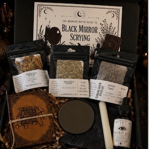 Könnte beinhalten: Eine schwarze Box mit einem Handbuch namens The Roaming Raven Guide to Black Mirror Scrying, drei Beuteln mit getrockneten Kräutern namens Sweet Dreams Tea, Mugwort und Lavender Flowers, einem braunen Lederbuch, einem schwarzen Spiegel, einer Kerze und Öl.