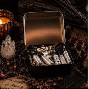 May include: A rectangular black metal tin containing several small glass vials with black screw caps. Each vial features a white paper label with black text and illustrations. The tin is open, revealing the vials nestled among shredded paper filler.