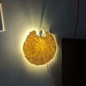 Puede incluir: Una lámpara de pared única con una pantalla circular y texturizada en un tono amarillo cálido. La pantalla parece estar hecha de material arrugado, iluminado desde atrás, proyectando un brillo suave. La lámpara está montada en una pared de color claro.