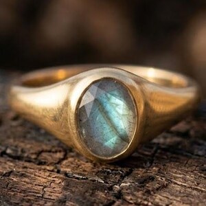 May include: A gold signet ring featuring a smooth, polished band and a central oval-shaped labradorite gemstone. The stone displays a characteristic iridescent blue-green flash within its faceted surface, set securely into the metal setting.