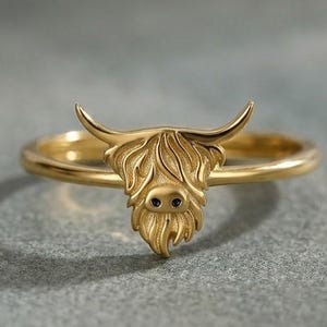 Puede incluir: Un anillo de tono dorado que presenta una cabeza de vaca Highland esculpida como punto focal central. El diseño de la vaca muestra patrones de pelaje detallados y texturizados, cuernos curvados y dos pequeños ojos circulares oscuros en un aro fino.