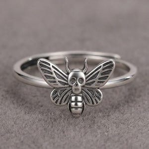 Può includere: Un anello in metallo color argento con un motivo centrale a forma di insetto, caratterizzato da una testa a forma di teschio e dettagliati motivi sulle ali. La fascia è sottile e circolare, con una finitura lucida e zone scure incise.