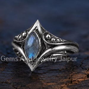 Puede incluir: Un anillo de plata con una piedra de labradorita en talla marquesa, engastada en un marco metálico geométrico puntiagudo con detalles de cuentas decorativas. La banda tiene un acabado pulido y oxidado que resalta el trabajo del metal.