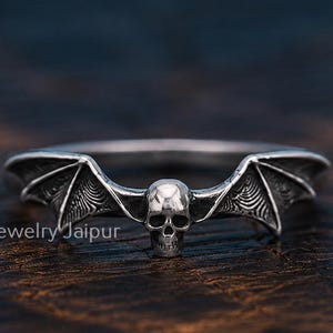 Puede incluir: Un anillo de plata con un diseño central de cráneo humano flanqueado por dos alas de murciélago detalladas que se extienden a lo largo de la banda. El metal tiene un acabado pulido con áreas oscurecidas para resaltar las texturas intrincadas del cráneo y las alas. El texto Gems And Jewelry Jaipur es visible en la esquina inferior izquierda.