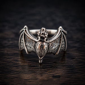 May include: A silver-toned ring featuring a detailed bat design with outstretched wings. The metal surface has an oxidized finish that highlights the textured lines and contours of the bat's body and wings, creating a distinct contrast.