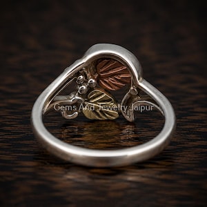Puede incluir: Un anillo de plata que presenta una secci&oacute;n superior decorativa con motivos de hojas en color cobre y oro, adem&aacute;s de peque&ntilde;os acentos esf&eacute;ricos. La banda es de plata lisa y pulida. El texto Gems And Jewelry Jaipur es visible en el anillo.