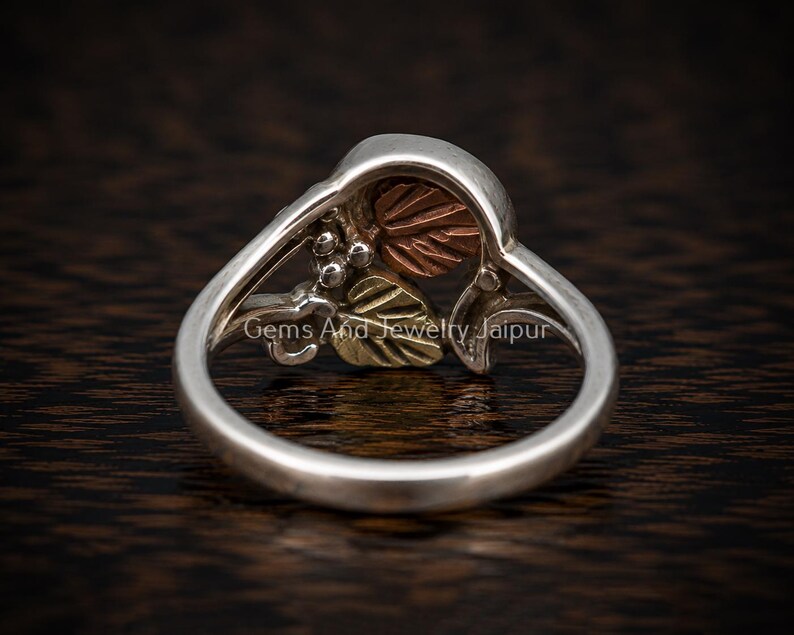 Puede incluir: Un anillo de plata que presenta una secci&oacute;n superior decorativa con motivos de hojas en color cobre y oro, adem&aacute;s de peque&ntilde;os acentos esf&eacute;ricos. La banda es de plata lisa y pulida. El texto Gems And Jewelry Jaipur es visible en el anillo.