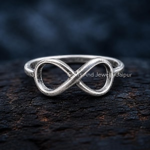 Puede incluir: Un anillo de metal plateado con un diseño de símbolo de infinito en el centro. La banda tiene un acabado texturizado y desgastado. El texto Gems And Jewelry Jaipur es visible en el centro del símbolo de infinito en una fuente clara.