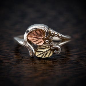 Puede incluir: Un anillo de plata con un dise&ntilde;o de hoja de oro rosa y una hoja de oro amarillo. La banda incluye peque&ntilde;as esferas de plata y detalles met&aacute;licos curvos. El texto Gems And Jewelry Jaipur es visible en el centro del anillo en letra clara.