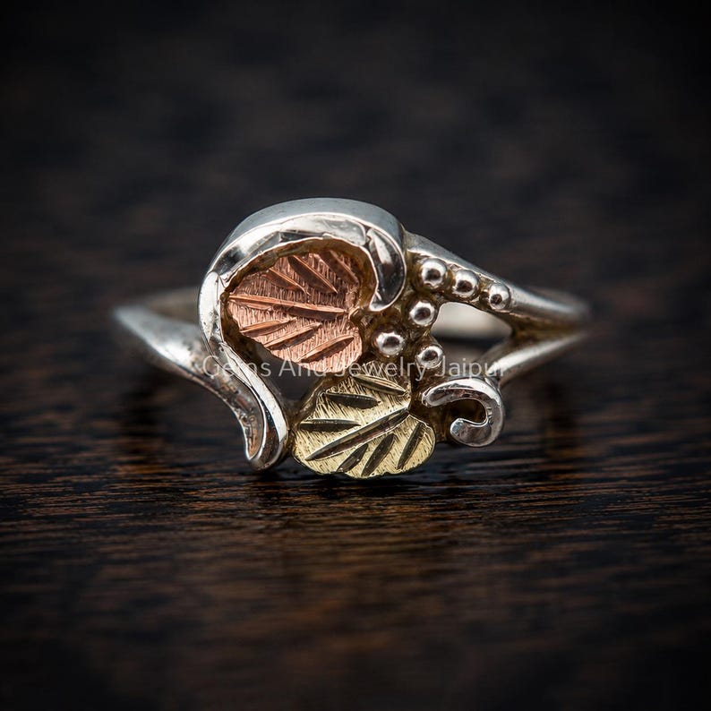 Puede incluir: Un anillo de plata con un dise&ntilde;o de hoja de oro rosa y una hoja de oro amarillo. La banda incluye peque&ntilde;as esferas de plata y detalles met&aacute;licos curvos. El texto Gems And Jewelry Jaipur es visible en el centro del anillo en letra clara.