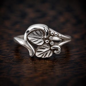Puede incluir: Un anillo de plata pulida que presenta un motivo decorativo de dos hojas texturizadas y un grupo de peque&ntilde;as cuentas esf&eacute;ricas. La banda es lisa y se estrecha hacia la parte posterior, con el dise&ntilde;o de hojas y cuentas centrado en el frente.