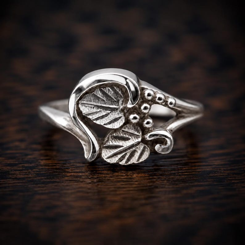 Puede incluir: Un anillo de plata pulida que presenta un motivo decorativo de dos hojas texturizadas y un grupo de peque&ntilde;as cuentas esf&eacute;ricas. La banda es lisa y se estrecha hacia la parte posterior, con el dise&ntilde;o de hojas y cuentas centrado en el frente.