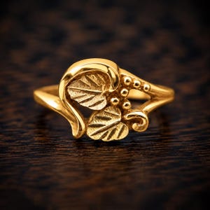 Puede incluir: Un anillo de oro con un dise&ntilde;o central de dos hojas texturizadas y un grupo de peque&ntilde;as cuentas esf&eacute;ricas. La banda es lisa y pulida, con el motivo de hojas integrado en el engaste, creando una apariencia bot&aacute;nica detallada y coherente.
