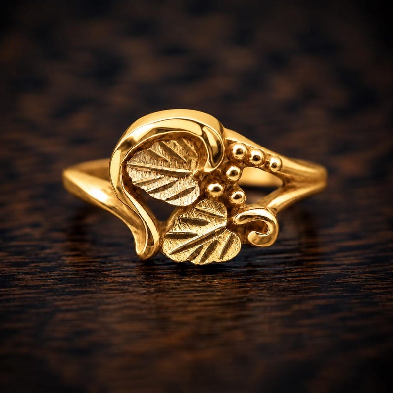 Puede incluir: Un anillo de oro con un dise&ntilde;o central de dos hojas texturizadas y un grupo de peque&ntilde;as cuentas esf&eacute;ricas. La banda es lisa y pulida, con el motivo de hojas integrado en el engaste, creando una apariencia bot&aacute;nica detallada y coherente.