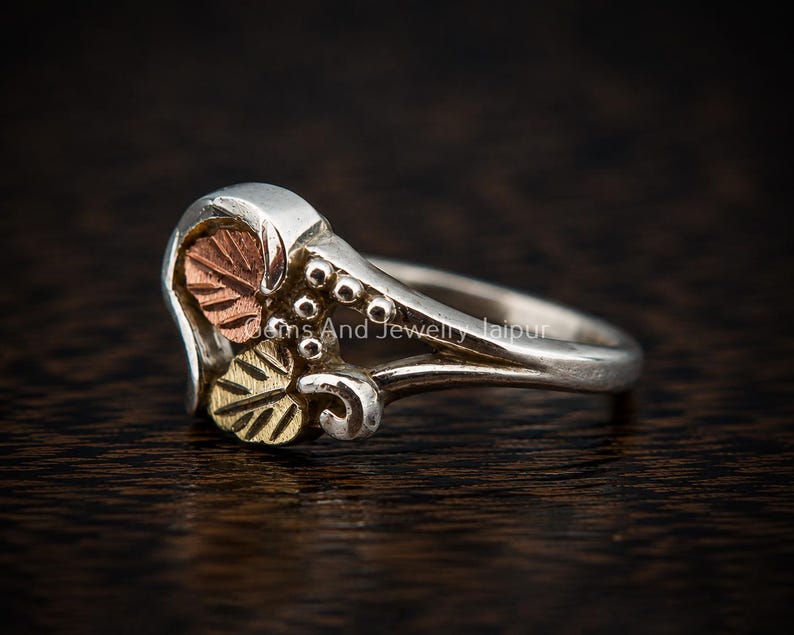 Puede incluir: Un anillo de plata con un motivo decorativo de hojas que incluye una hoja de tono cobrizo y una hoja de tono dorado, acentuado por peque&ntilde;as esferas de plata. El texto Gems And Jewelry Jaipur es visible en la superficie del anillo.