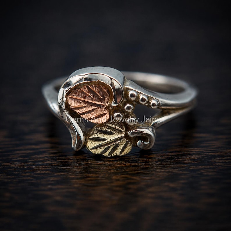 Puede incluir: Un anillo de plata con un dise&ntilde;o decorativo de dos hojas, una en oro rosa y otra en oro amarillo, acentuado con peque&ntilde;as esferas de plata. La banda es de plata pulida e incluye el texto Gems Jewelry Jaipur grabado en la superficie.