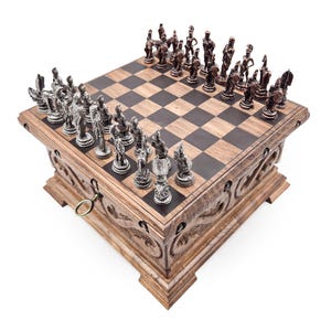 Puede incluir: Un juego de ajedrez de madera con detalles tallados. El tablero presenta un patrón de cuadros oscuros y claros. Las piezas de ajedrez son de color plateado y bronce, que representan figuras. El juego incluye un cajón con una llave. Un juego de estrategia clásico.