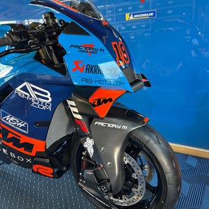 Può includere: Una moto KTM blu e arancione con il testo "FACTORY M CARBON WORKS" e "AKRAPOVIC" visibili. La moto ha uno pneumatico nero e un parabrezza trasparente. Il numero 19 è visualizzato sulla parte anteriore.