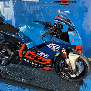 Könnte beinhalten: Ein blau-oranges KTM-Motorrad mit den Texten "RACE BOX" und "AB-MOTO.COM". Das Motorrad hat einen schwarzen Sitz und eine klare Windschutzscheibe. Das Motorrad steht auf einer Hebebühne.