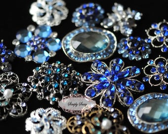May include: A collection of various blue and silver brooches and pins. The brooches feature different designs, including floral and geometric shapes, and are adorned with blue gemstones and crystals. The brooches are displayed on a black surface.