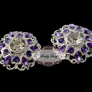 Puede incluir: Dos broches de plata con un diseño de filigrana y un gran cristal de roca transparente en el centro. Los broches están rodeados de cristales de roca morados más pequeños.