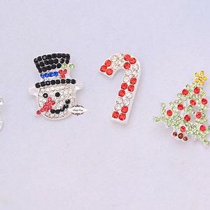 May include: Five Christmas-themed rhinestone embellishments: a red Santa hat, a snowman with a black top hat, a red and white candy cane, a green and red Christmas tree, and a red stocking.