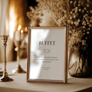 Könnte beinhalten: Ein goldgerahmtes Schild mit dem Wort "BUFFET" in schwarzer Schrift. Das Schild enthält deutschen Text und eine Grafik von Teller und Gabel. Ein goldener Kerzenständer und ein Blumenarrangement befinden sich im Hintergrund.