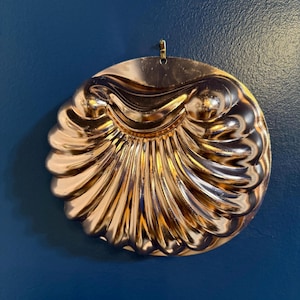 May include: A copper-coloured, round decorative mould with a seashell design. The mould has a hanging loop at the top and is made of shiny metal. The design features curved ridges and a scalloped edge.