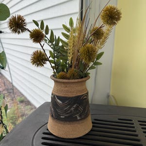 May include: A small, handcrafted ceramic vase with a tan base and a dark, swirled design around the middle. The vase holds a collection of dried, spiky seed heads, green leaves, and wheat stalks, creating a rustic, natural arrangement.