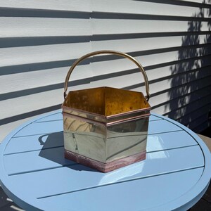 May include: A vintage brass bucket with a copper trim and a curved brass handle. The bucket has a square shape and is sitting on a light blue table. The bucket is made of metal and has a shiny finish.