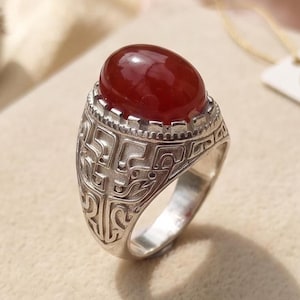 May include: A silver ring featuring a large, smooth, oval-shaped deep red stone set in a bezel. The band is crafted from silver metal with intricate, recessed geometric patterns and scrollwork detailing that wraps around the sides of the ring.