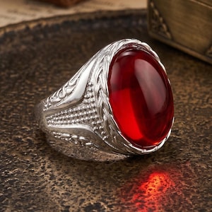 Può includere: Un anello d'argento con una gemma ovale rosso scuro. L'anello presenta un design ornato e strutturato attorno alla pietra e sulla fascia. La gemma è lucidata e riflette la luce. Un gioiello.