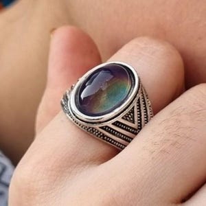 以下が含まれることがあります： 紫、青、緑の虹色のグラデーションを示す楕円形のムードストーンをあしらったシルバー調のリング。金属製のバンドには、刻印された線と小さなドットのディテールが施された、質感のある幾何学模様がデザインされています。
