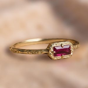 Puede incluir: Un anillo de oro con una gema rectangular de color rojo intenso. El anillo tiene un diseño de filigrana ornamentado alrededor de la gema y en la banda. El anillo está sobre un fondo suave y borroso.