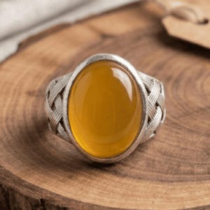 May include: A silver ring featuring a large, smooth, oval-shaped yellow gemstone set in the center. The band of the ring displays a detailed, textured woven or braided metal pattern that extends from the central stone setting around the sides.