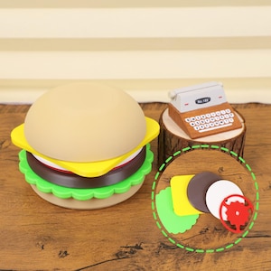 Puede incluir: Un juguete caprichoso con forma de hamburguesa con pan tostado, queso amarillo, una hamburguesa marrón y lechuga verde. Una máquina de escribir en miniatura sobre una base de madera.