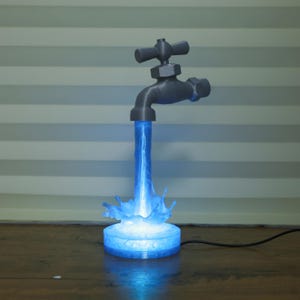 May include: A 3D-printed lamp shaped like a faucet, with a stream of blue light emanating from the spout. The light forms a splash at the base, and the faucet is gray. The lamp is on a wooden surface.