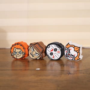 May include: Four colorful shoe charms featuring iconic horror movie characters. The charms include Chucky with orange hair, Freddy Krueger with a brown hat, Jason Voorhees' hockey mask, and Pennywise with orange hair.
