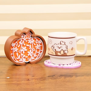 May include: A brown pumpkin-shaped coaster with a white floral design and orange backing sits next to a white mug with a cartoon character and a purple coaster. The mug has a brown band around it.
