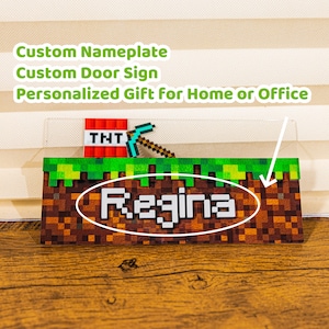 May include: A custom nameplate with the name "Regina" in white block letters. The design features a pixelated Minecraft theme with a TNT block, pickaxe, and a grass and dirt block design. Text above reads "Custom Nameplate, Custom Door Sign, Personalized Gift for Home or Office."