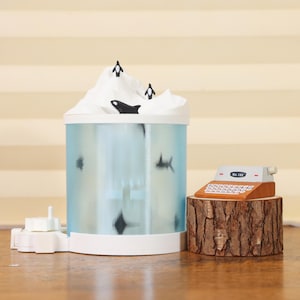 以下が含まれることがあります： ペンギンとシャチのフィギュアが特徴の、山型の蓋が付いた装飾的な水族館。 「No. 155」の文字が入ったミニチュア木製レジが、木の幹の台座に置かれています。 水族館は淡い青色です。