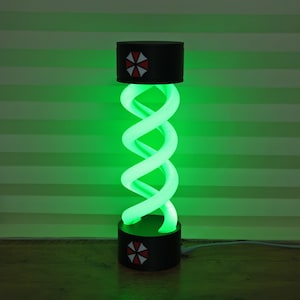 May include: A green glowing lamp with a spiral design. The lamp has a black base and top with a red and white emblem. The lamp is plugged in with a white cord. The background is a light green.