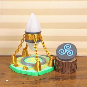 May include: A fantasy-themed lamp with a white, faceted light source suspended by gold chains. The lamp sits on a green base with gold accents. A grey stone with a blue spiral design rests on a wood slice.