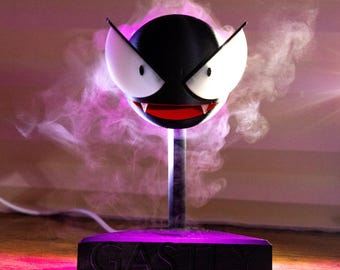 May include: A black and white Gastly figure with a red mouth and sharp teeth, mounted on a stand. The base of the stand is black with the word "GASTLY" in raised letters. The figure is surrounded by a purple mist.