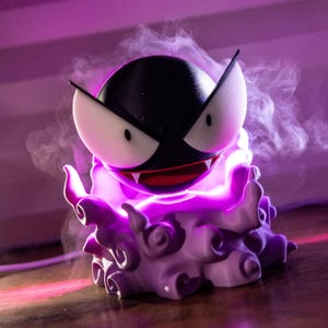 May include: A purple and black character lamp with a smiling face, sharp teeth, and pointed ears. The lamp emits a purple glow and has a smoky effect around it. The lamp is a stylized representation of a cartoon character.