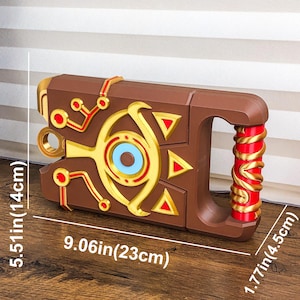 Pode incluir: Um adere&ccedil;o castanho com detalhes dourados e vermelhos, com uma pega e padr&otilde;es geom&eacute;tricos. O adere&ccedil;o mede 23 cm de comprimento, 14 cm de altura e 4,5 cm de largura. O design inclui um s&iacute;mbolo central em forma de olho.